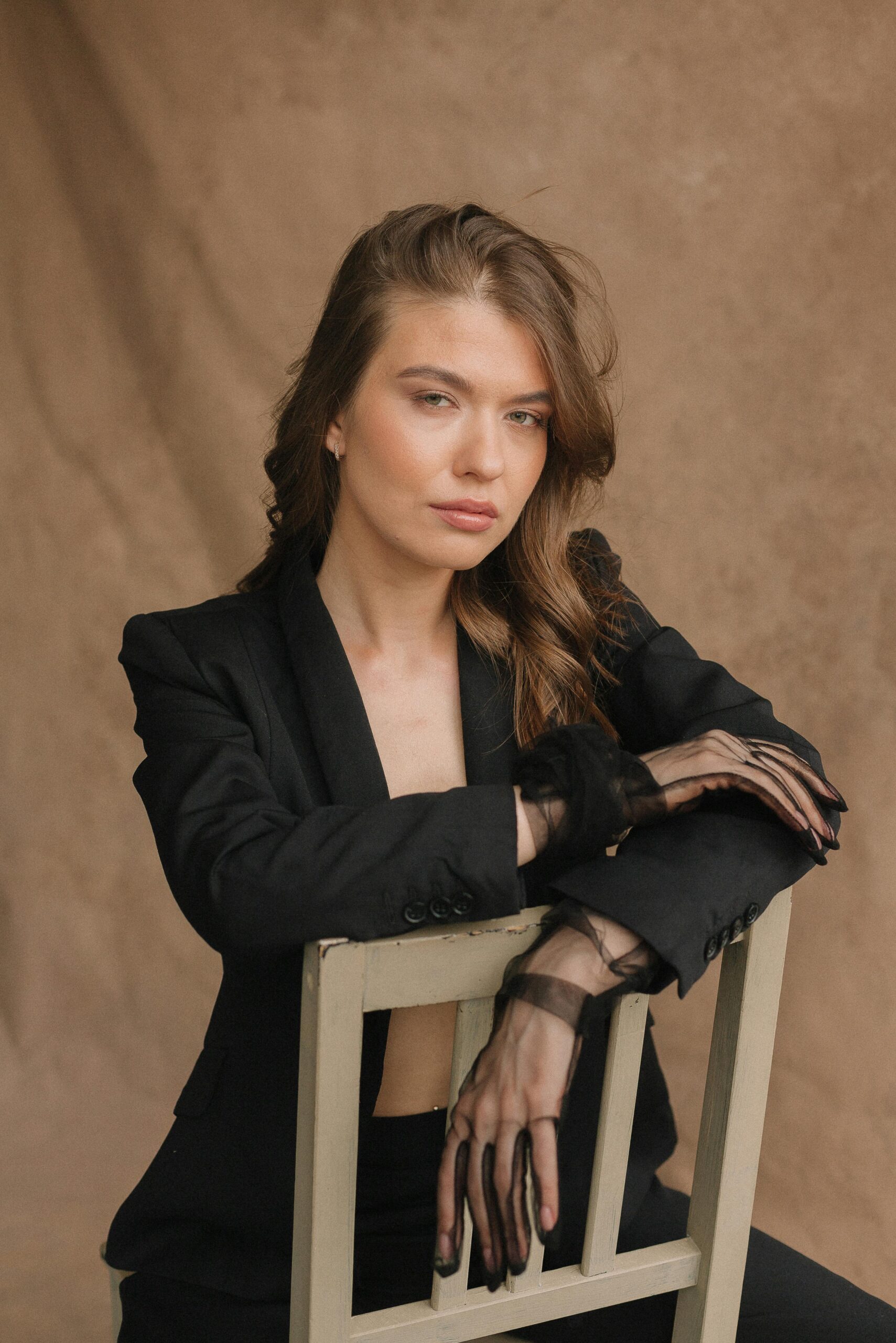 Elegant woman with long wavy hair seated, wearing black clothing and sheer gloves in a studio setting.